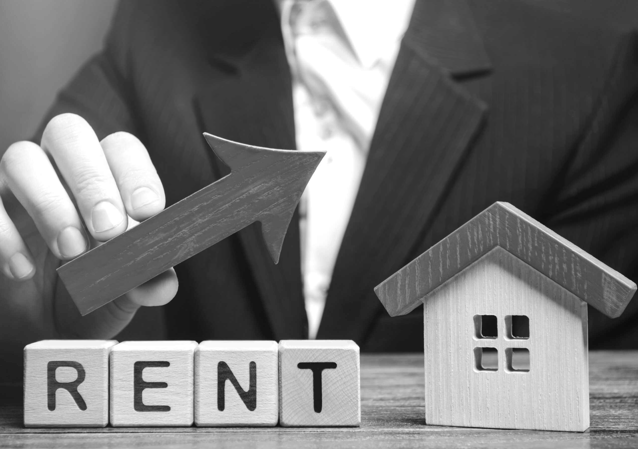 Изображение на човек с костюм, държащ стрелка. Пред него има дървена къщичка и надпис RENT, изобразяваща покачването на дългосрочните наеми.